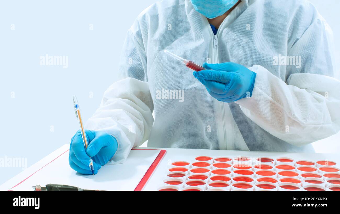 Da vicino al medico con guanti blu che scrivono su un foglio bianco di carta, tenendo una siringa con sangue in mano, seduto accanto ad un gruppo di cliniche Foto Stock