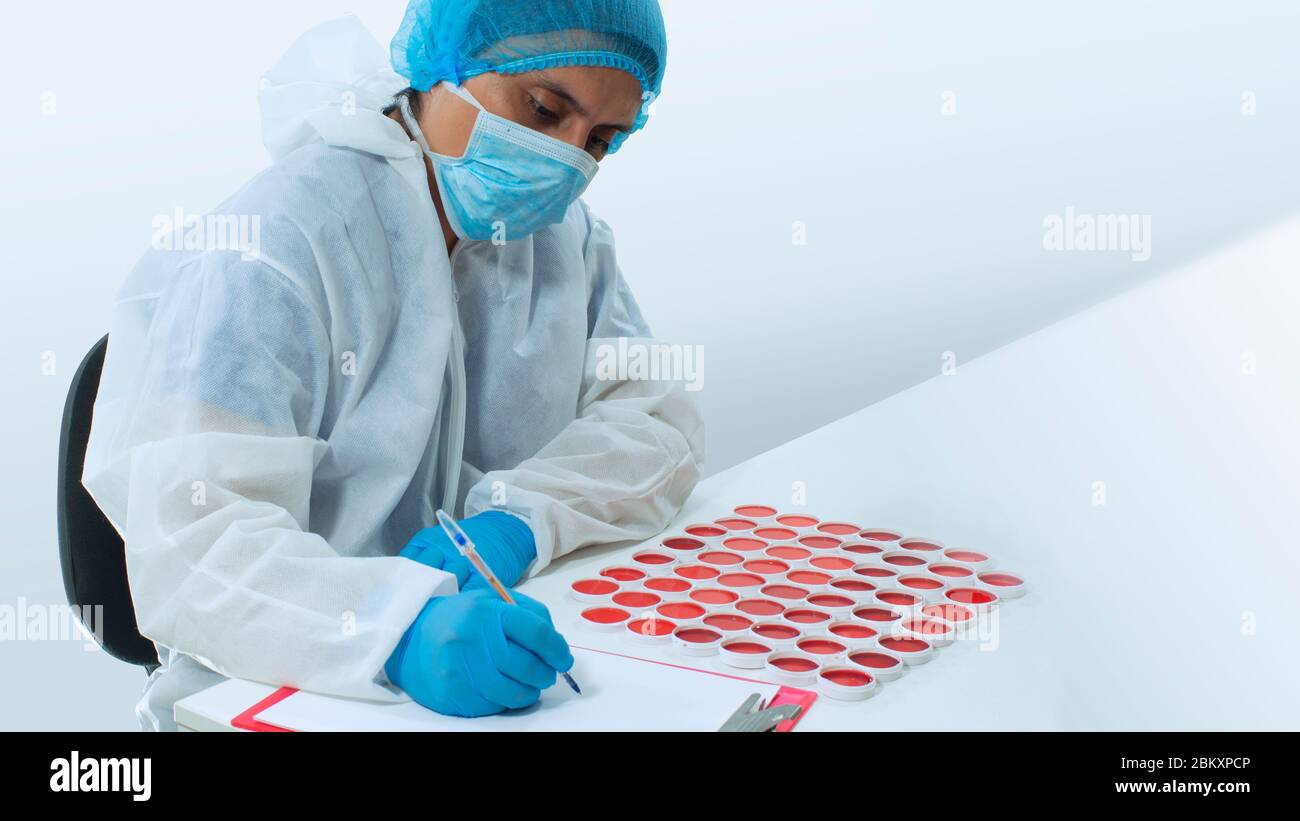 Medico con maschera e guanti blu che scrivono su un foglio bianco di carta accanto ad un gruppo di campioni clinici di liquido rosso in contenitori bianchi rotondi Foto Stock