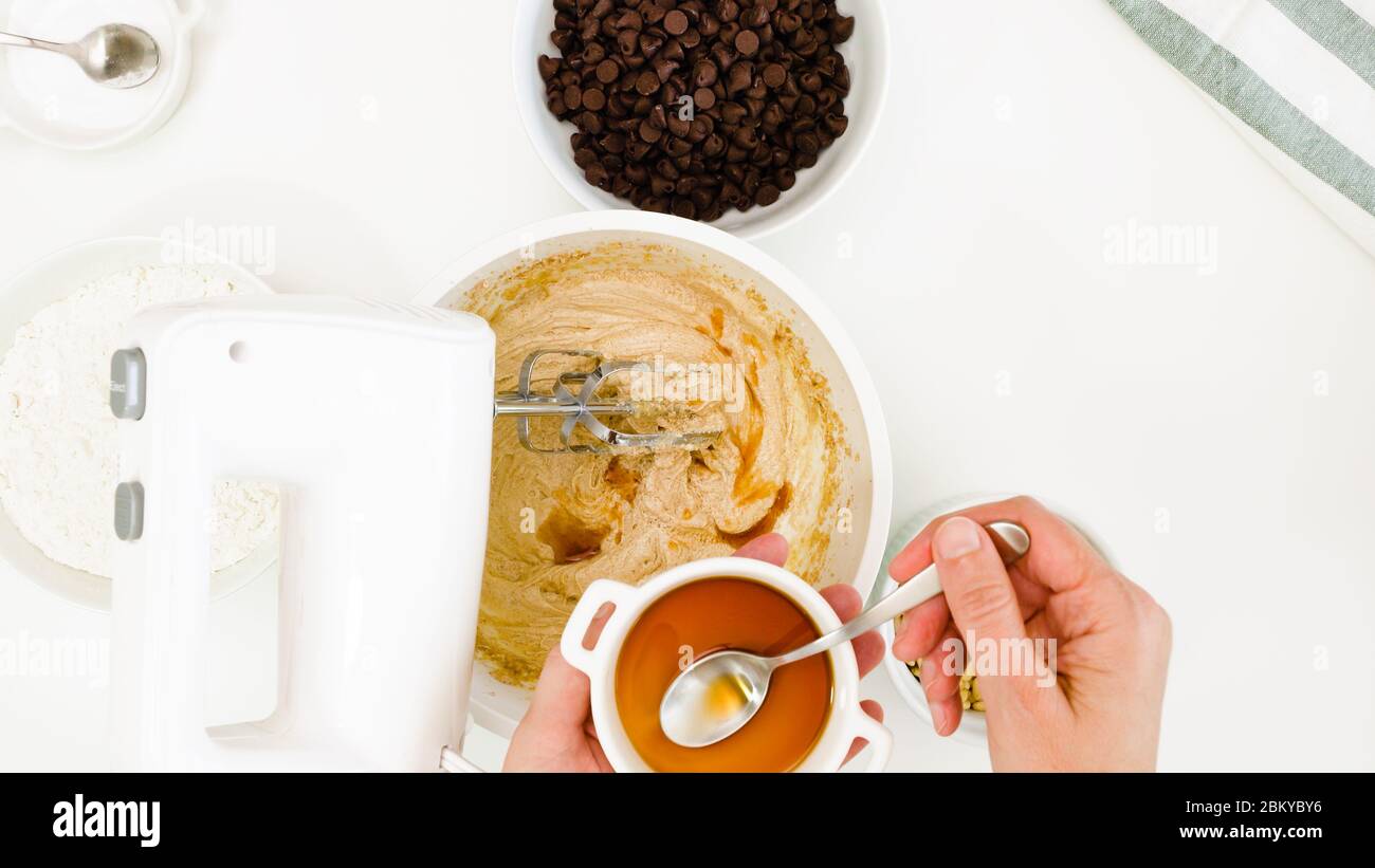 Ricetta dei biscotti del chip del cioccolato. Chef aggiunge pochi cucchiaini di estratto di vaniglia in una pastella di biscotti, primo piano su sfondo bianco, vista dall'alto Foto Stock