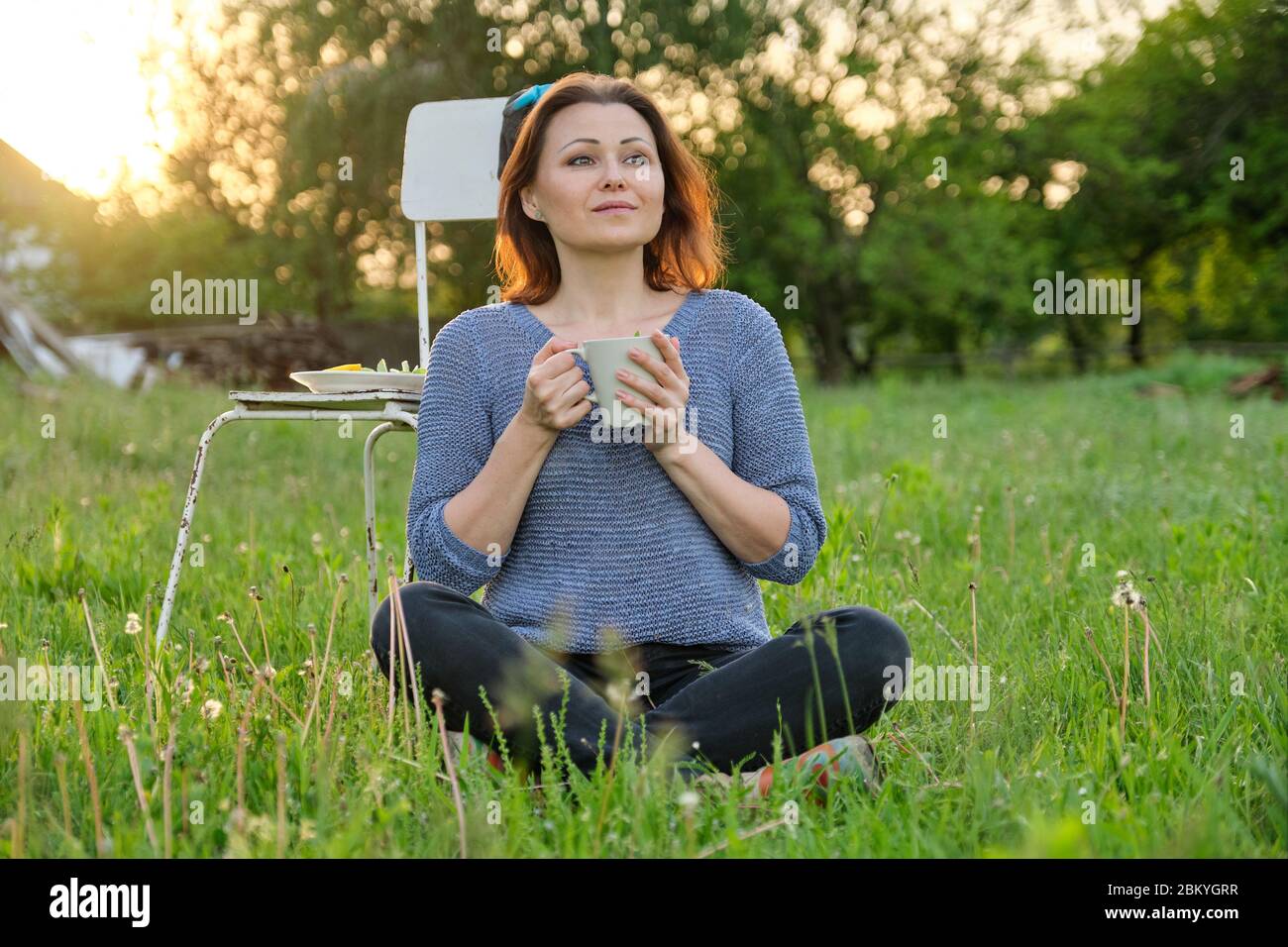 Bella donna di mezza età seduta su erba in giardino, rilassante al tramonto Foto Stock