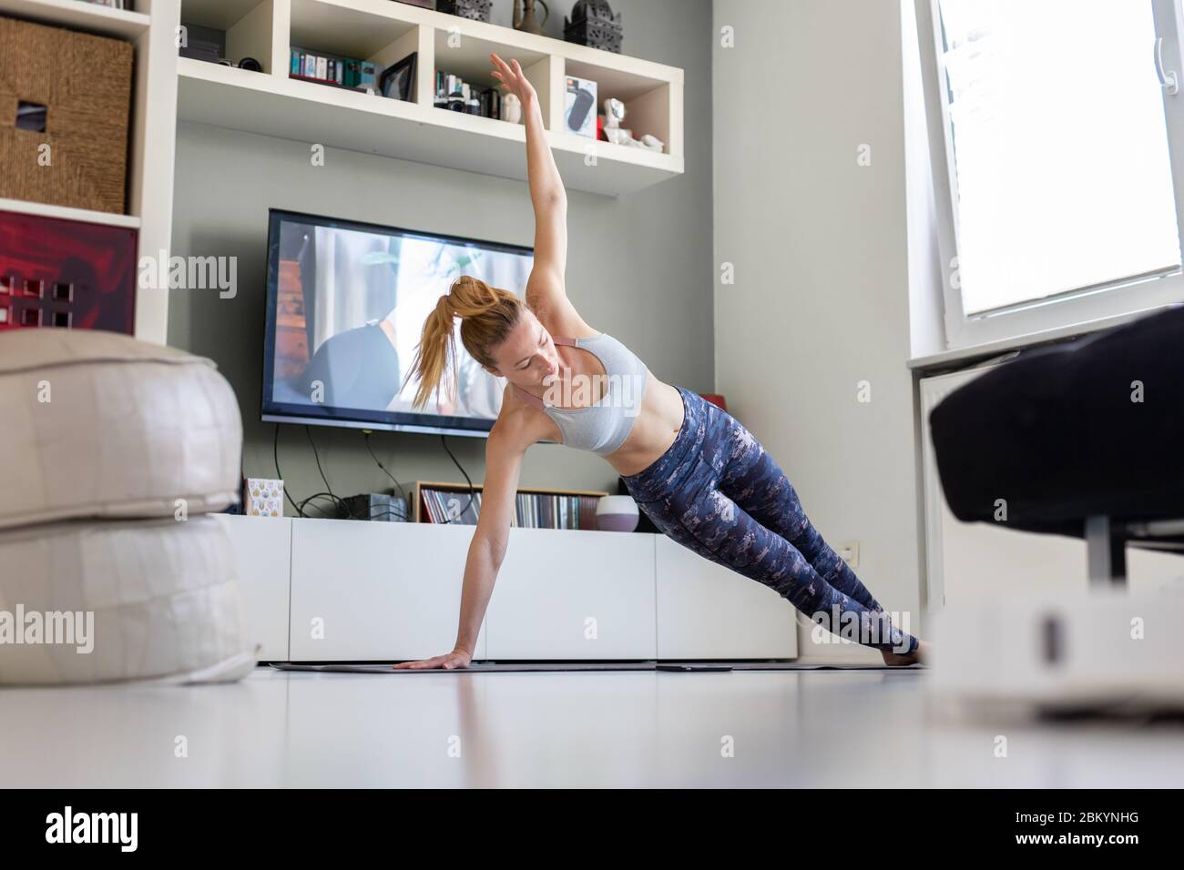 Donna sportiva attraente che si allena a casa, facendo esercizio di pilates davanti alla televisione nel suo soggiorno. Distanza sociale. Rimanere sani e. Foto Stock