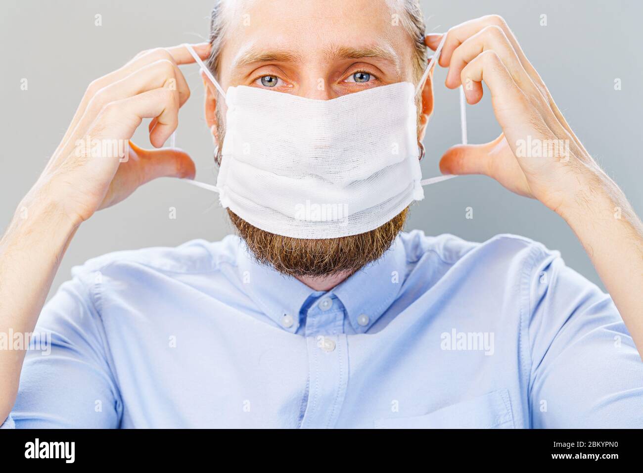 L'uomo bianco bearded in camicia blu mette su una maschera di inquinamento dell'aria e di coronavirus Covid19, guardando la macchina fotografica. Studio girato su sfondo grigio Foto Stock