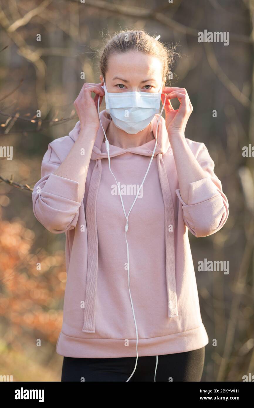 Ritratto della donna sportiva caucasica che indossa la maschera facciale di protezione medica mentre cammina nel parco, rilassandosi e ascoltando la musica. Corona virus, o Covid Foto Stock