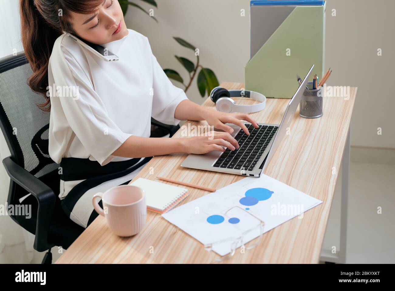 Fiducioso donna asiatica parlando sul suo telefono cellulare e utilizzando computer portatile mentre è seduto alla scrivania e lavora da casa. Home office. Foto Stock