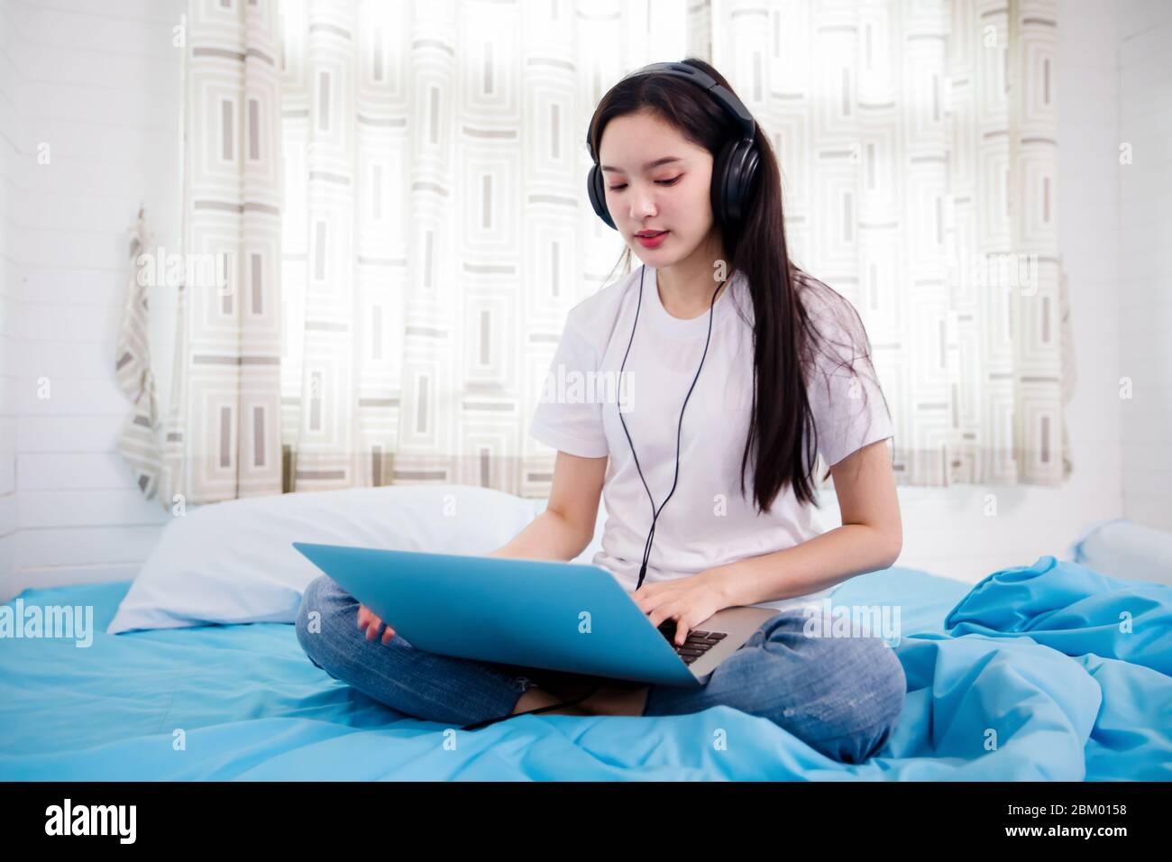 Giovane donna felice asiatica indossa cuffie e ascoltare musica canzone e guardare il sito web per lo shopping online dal notebook a letto a casa Foto Stock