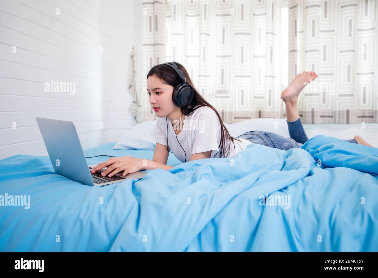 Giovane donna felice asiatica indossa cuffie e ascoltare musica canzone e guardare il sito web per lo shopping online dal notebook a letto a casa Foto Stock