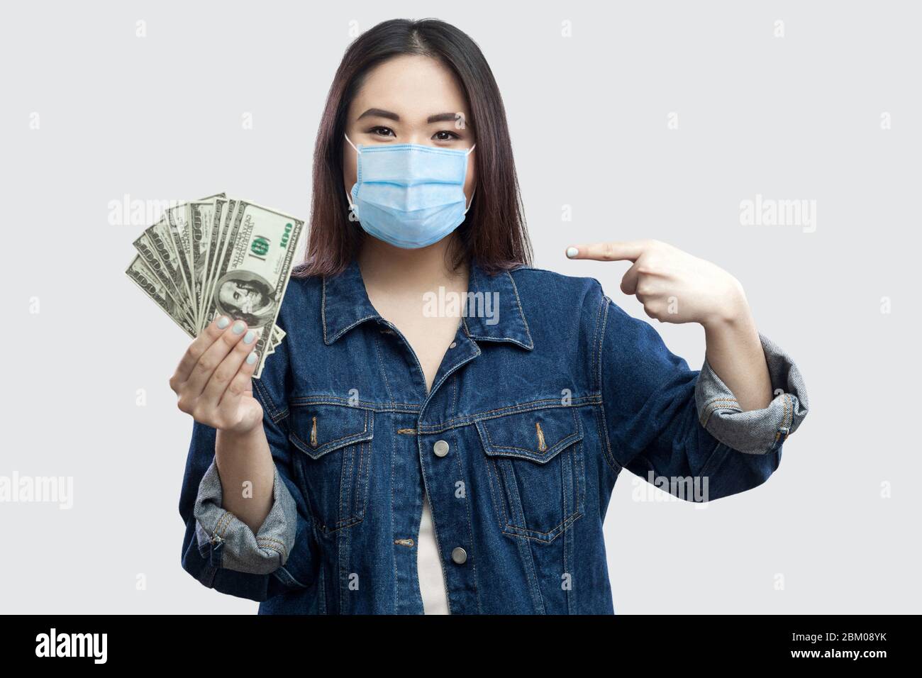 Felice soddisfatto bella giovane donna d'affari con maschera medica in blu giacca denim in piedi, tenendo molti dollari, dito di puntamento e guardando ca Foto Stock