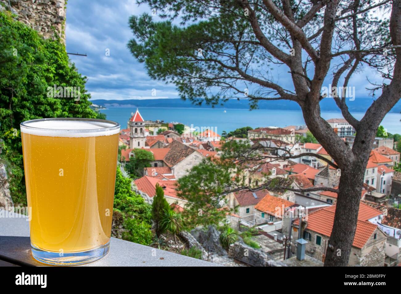 Bicchiere di birra leggera con vista sulla città mediterranea, Omis, Croazia. Una pausa con un bicchiere di birra e vista sui tetti rossi e sul mare blu. Foto Stock