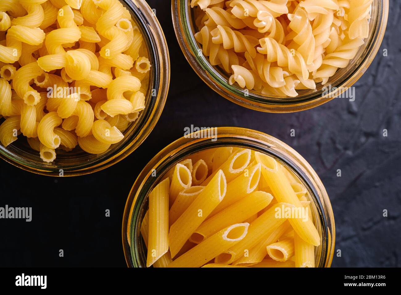 Tre vasetti di vetro con varietà di pasta di grano dorato crudo su sfondo nero scuro testurizzato, macro vista dall'alto Foto Stock