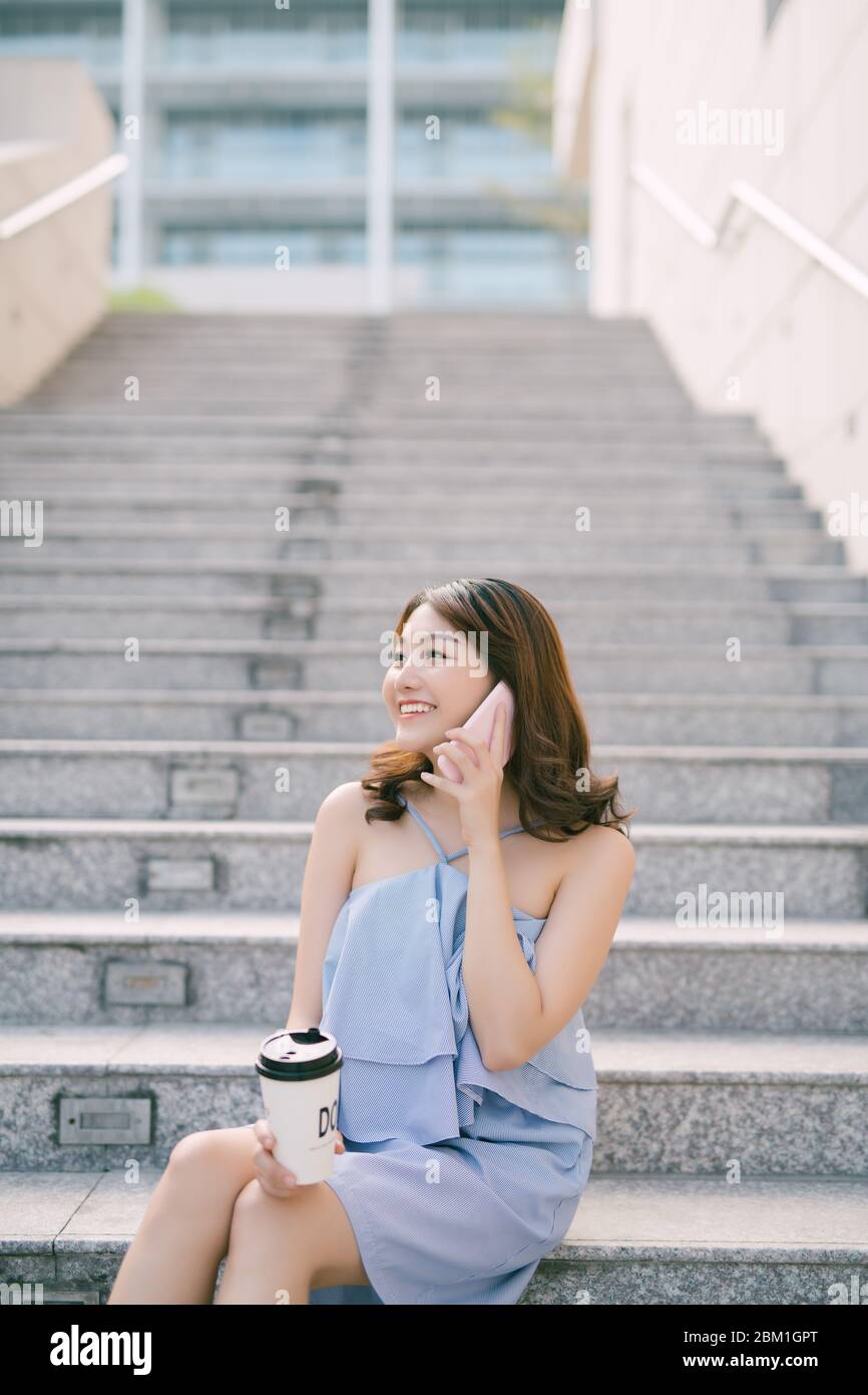 Allegro giovane ragazza asiatica parlando al telefono mentre è seduto alla scala esterna. Stile di vita urbano della ragazza. Foto Stock