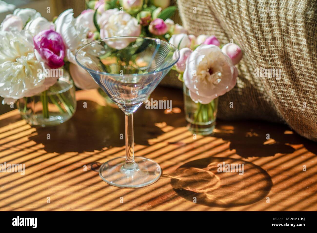 Festosa vita rustica con vetro vuoto vetro trasparente circondato da fiori bianchi su sfondo tessile su tavolo di legno. Ombra diagonale a righe di t Foto Stock