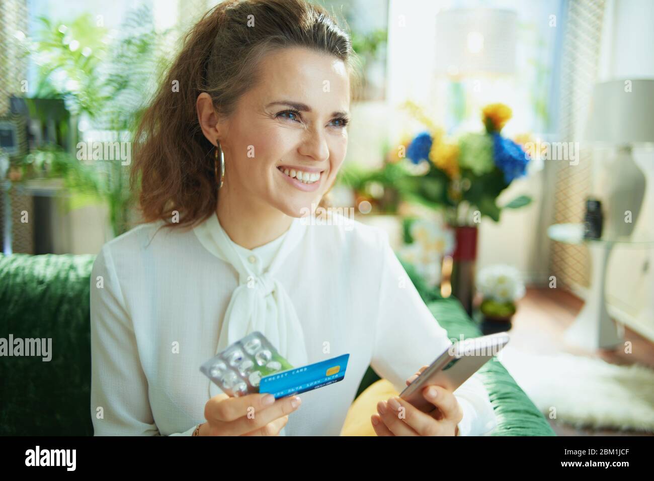 sorridente elegante femmina in blusa bianca con carta di credito e blister vuoto confezione di pillole utilizzando online farmacia negozio su uno smartphone nel moderno hous Foto Stock