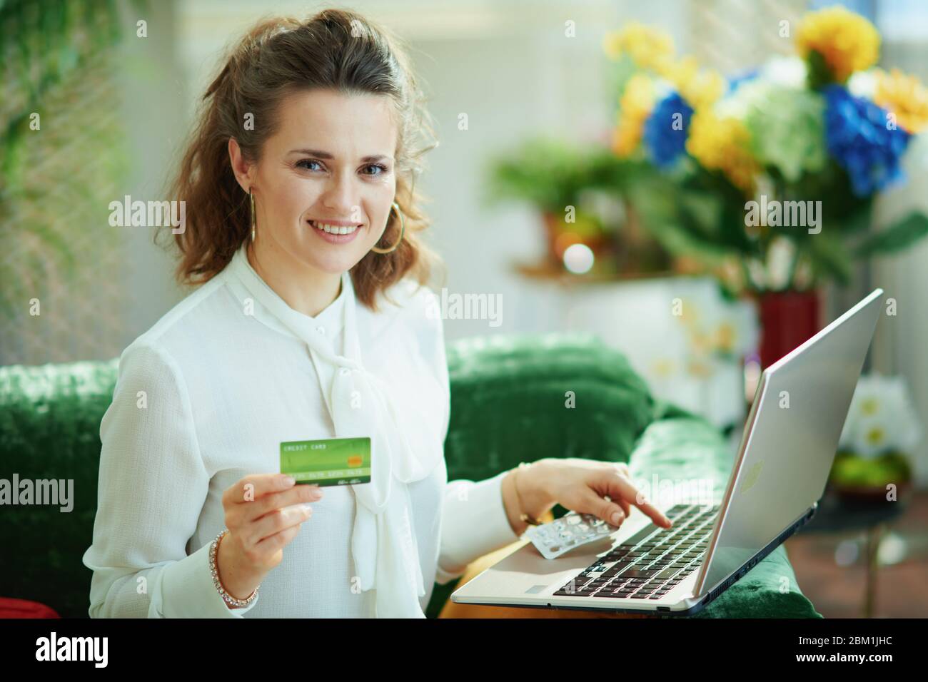 donna di media età sorridente in blusa bianca con carta di credito e blister vuoto confezione di pillole acquistare farmacia su internet utilizzando il laptop in moder Foto Stock