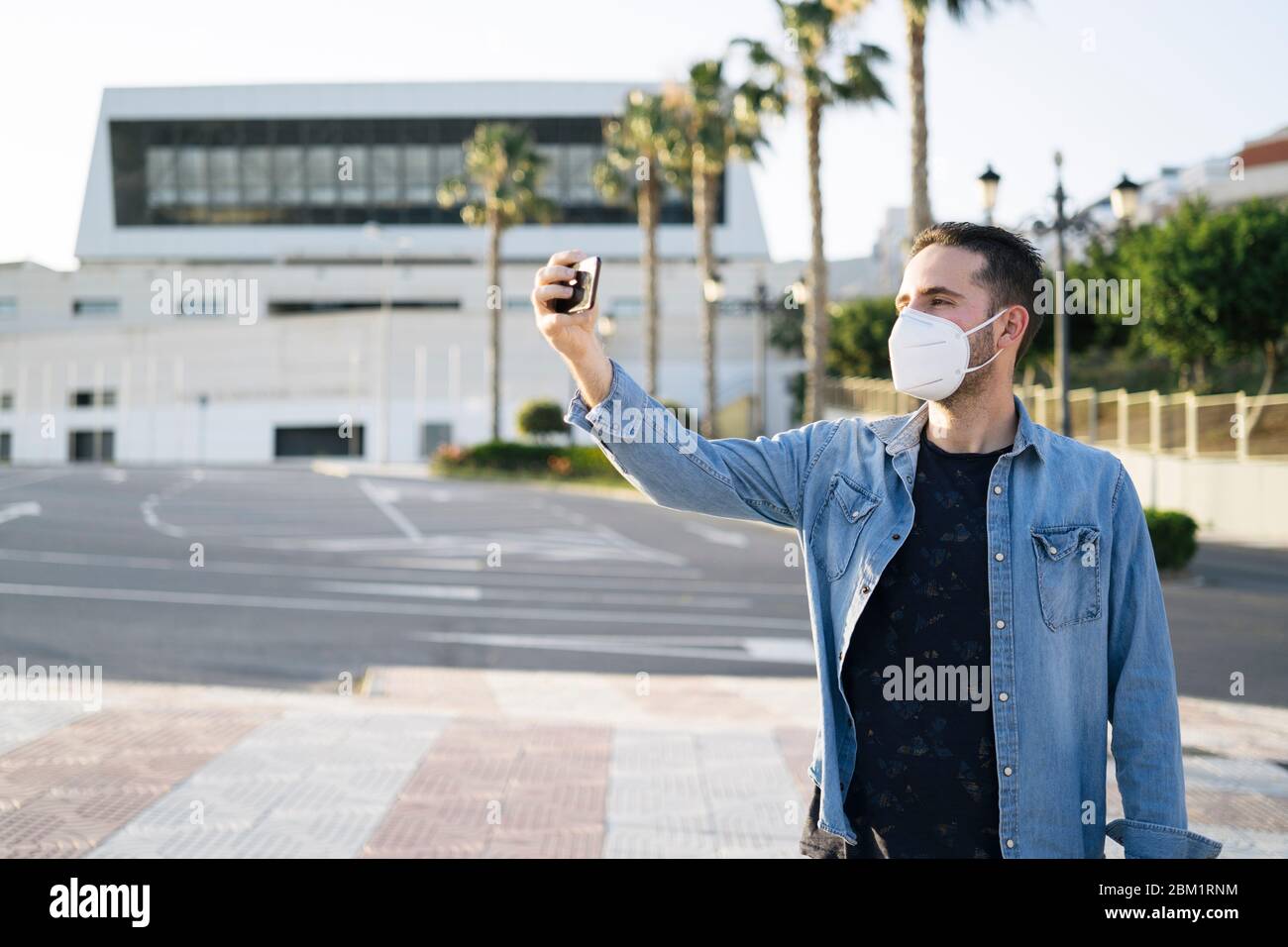 Giovane uomo che indossa una maschera medica facendo selfie con il suo smartphone. Un uomo in una maschera protettiva è protetto dal virus in strada. Foto Stock