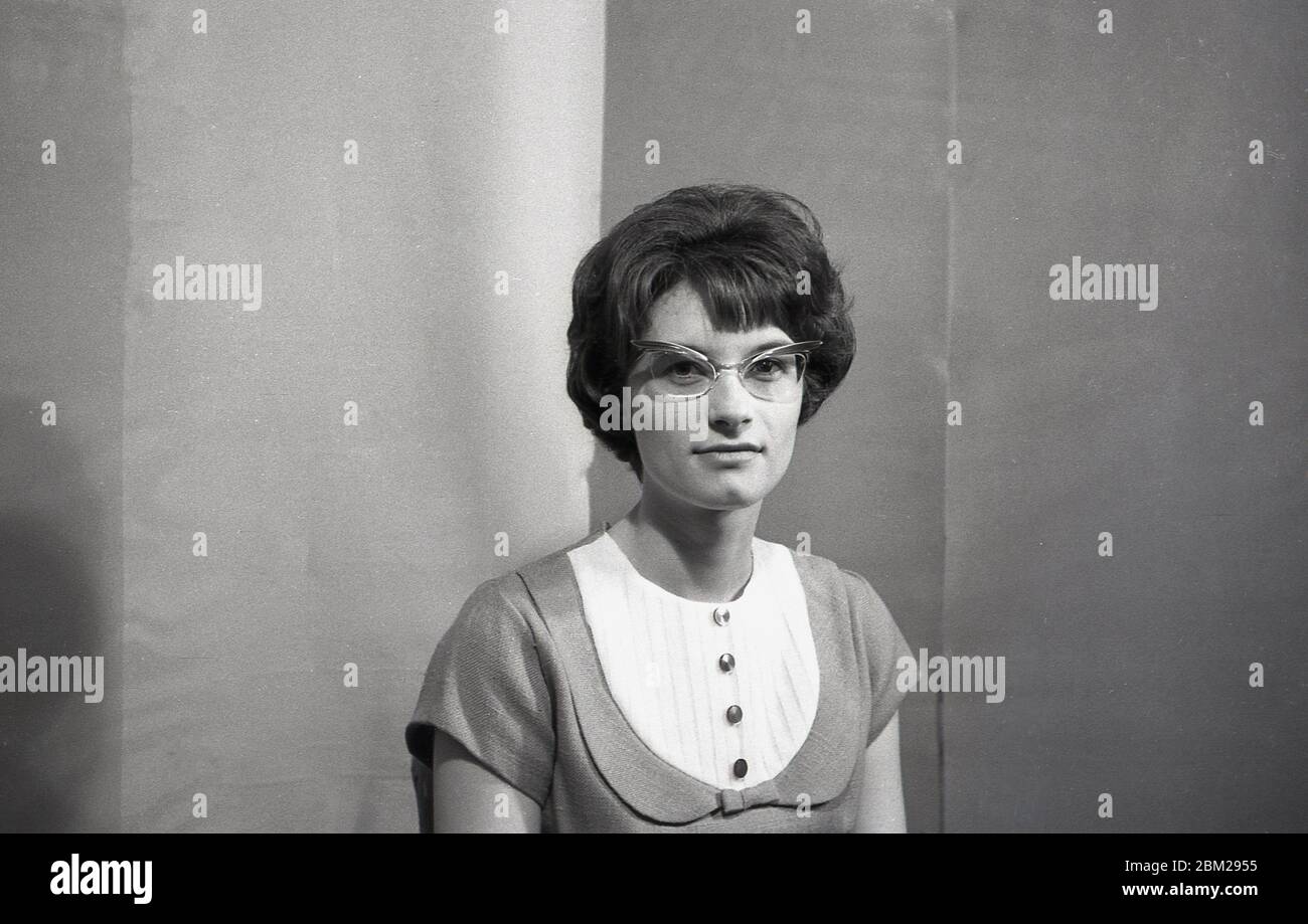 1961, ritratto storico e coinvolgente di una giovane donna con un top, acconciature e occhiali distintivi dell'epoca seduta in uno studio, Inghilterra, Regno Unito. Foto Stock