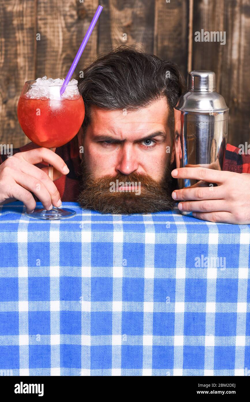 Barman o hipster tiene un cocktail con ghiaccio e paglia. Barman con barba  e faccia rigida mette la testa sulla tovaglia a scacchi. L'uomo tiene il  vetro e lo shaker su sfondo