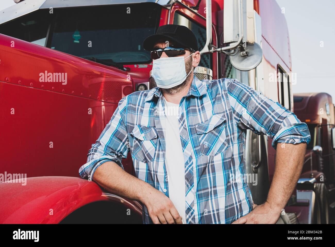 il conducente professionista del semi-camion sul lavoro in abbigliamento casual indossa la maschera facciale medica di sicurezza. Un autotreno dall'aspetto sicuro si trova accanto al carro di perforazione rosso grande Foto Stock