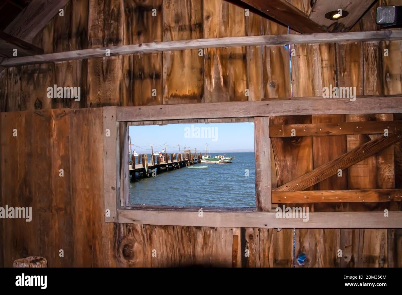 Vista blu dell'oceano con un molo e una barca da una finestra all'interno di un vecchio shack di legno Foto Stock