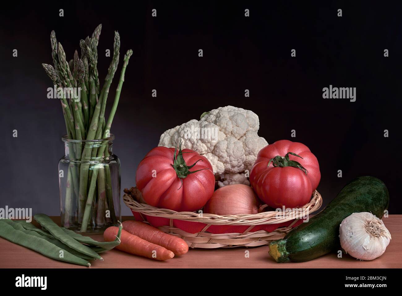 Ancora vita di frutta, verdura e un cesto rustico di materiali naturali, vaso di vetro trasparente con asparagi selvatici su sfondo scuro. Foto Stock