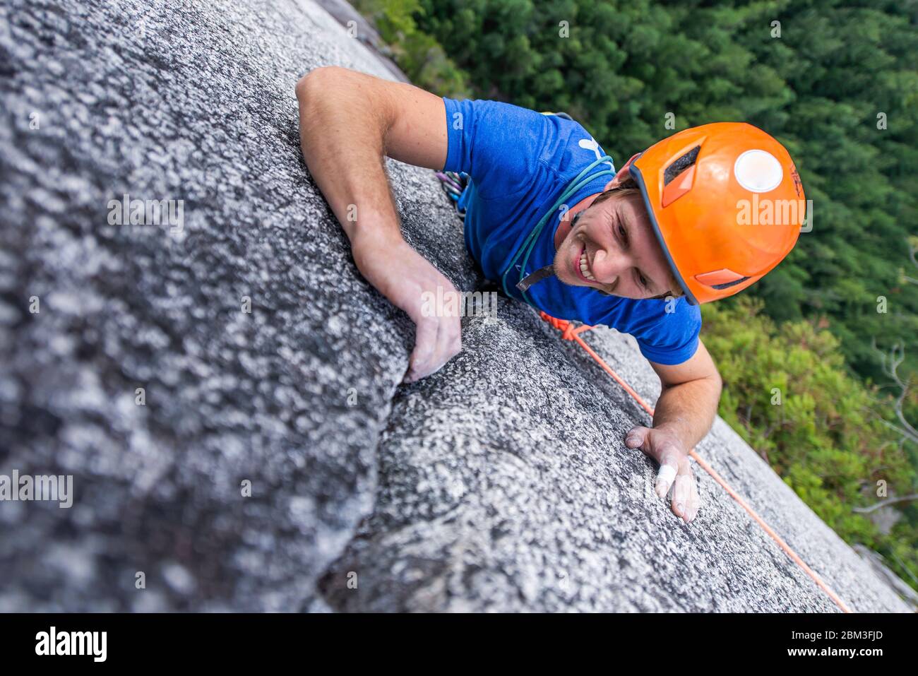 arrampicatore guardando in su e ridendo mentre arrampica su roccia con il casco Capo Foto Stock