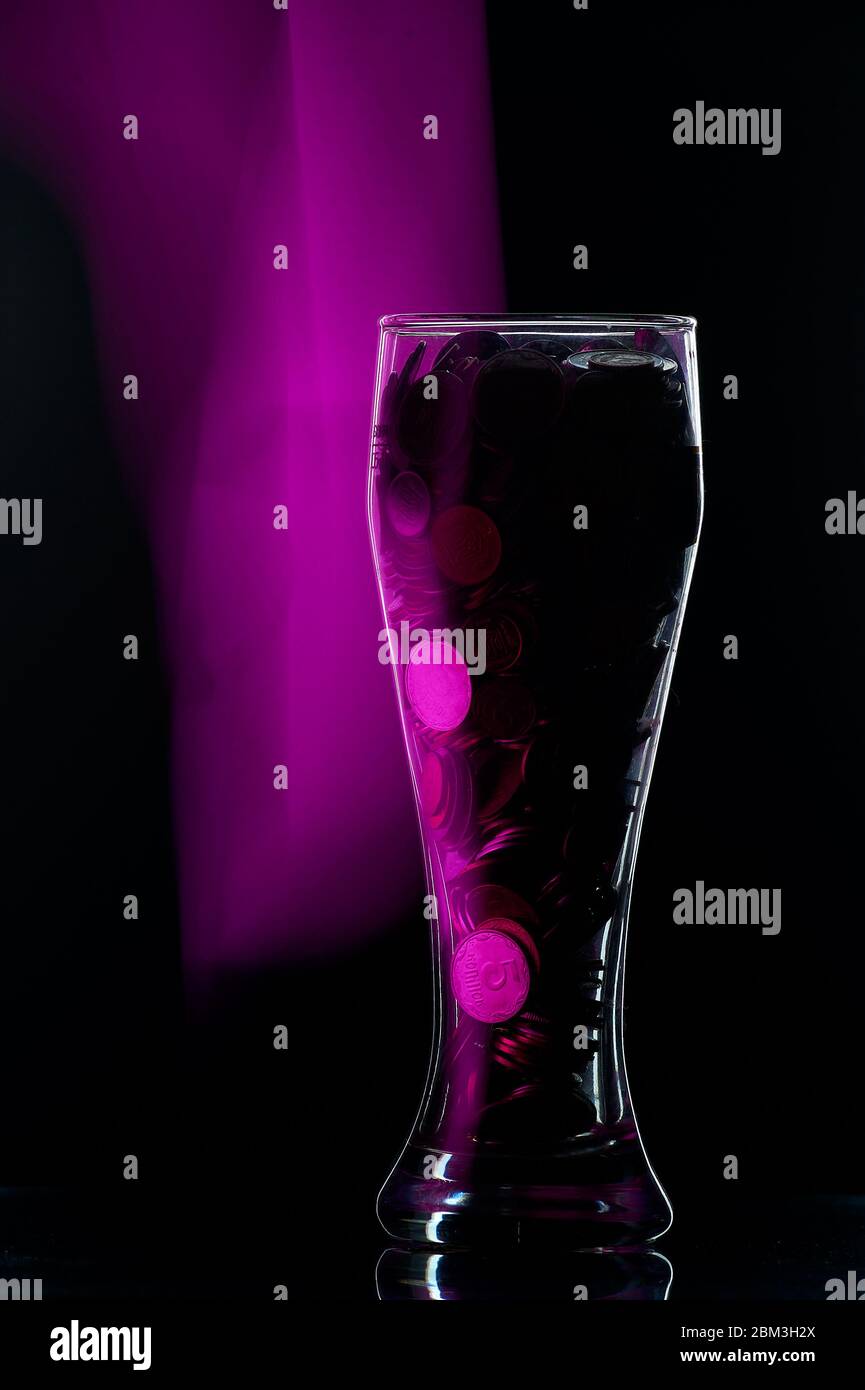 bicchiere di birra pieno di monete su sfondo nero con retroilluminazione Foto Stock