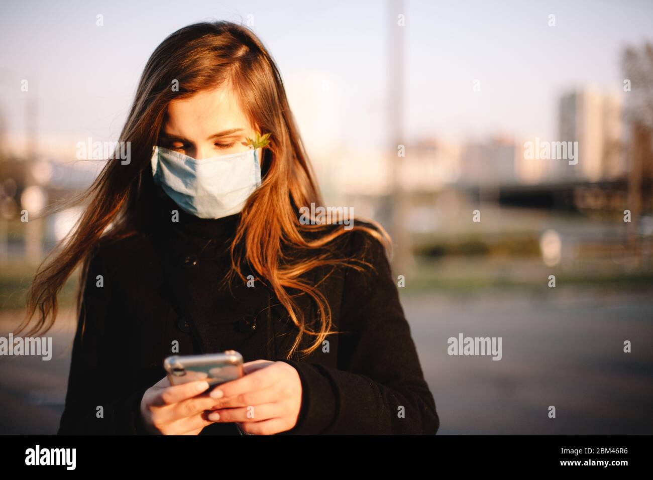 Ritratto di ragazza adolescente triste utilizzando smartphone indossando maschera medica viso protettivo in piedi all'aperto sulla strada della città Foto Stock