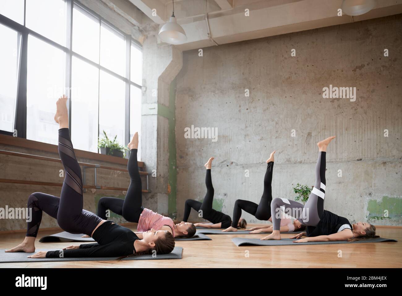 Vista laterale di un gruppo di ragazze sportive che allenano lo yoga nella sala fitness. Donne giovani in forma che si allungano, sdraiati su tappetini con gamba in palestra con grandi finestre. Interno loft. Concetto di sport. Foto Stock