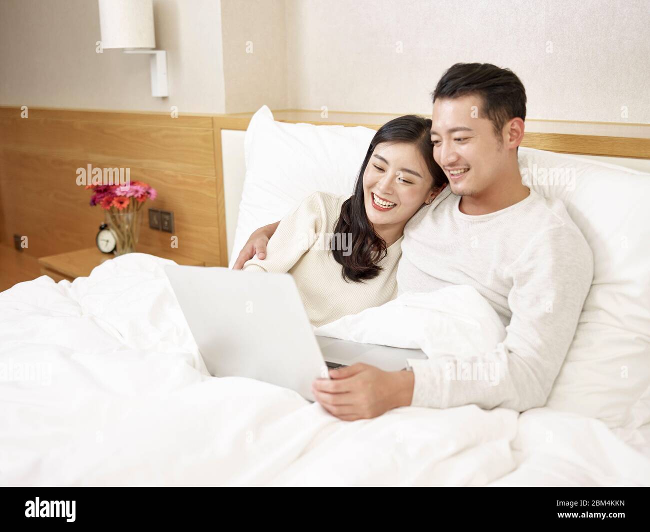 giovane coppia asiatica seduta a letto godendo un film utilizzando il computer portatile felice e sorridente Foto Stock