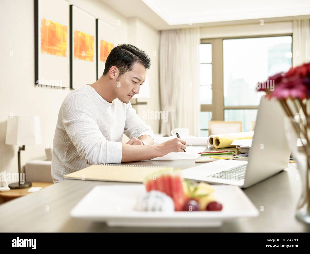 giovane asiatico uomo design professionale lavorare da casa seduta al banco della cucina guardando un disegno (grafica in background alterata digitalmente) Foto Stock