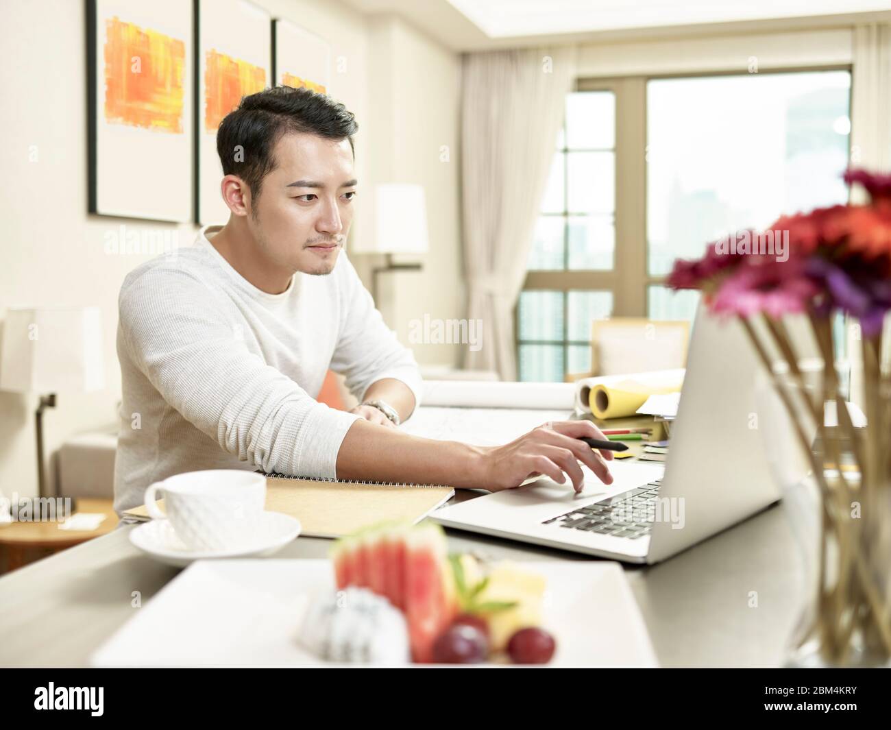 giovane uomo d'affari asiatico che lavora da casa seduto al bancone della cucina guardando il computer portatile (grafica in background modificata digitalmente) Foto Stock