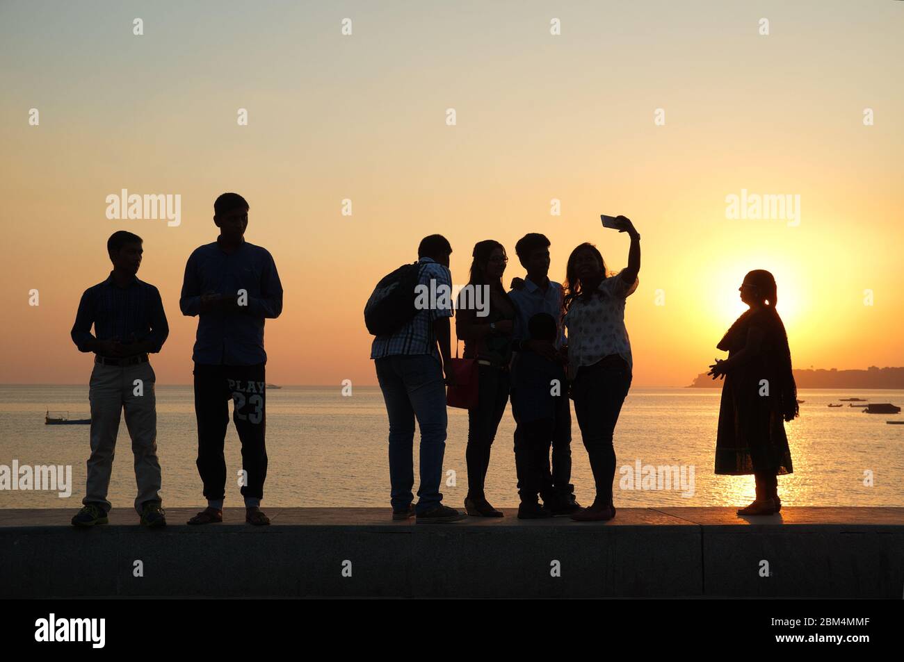 Un gruppo di giovani al tramonto che si erge sulla parete di argine di Marine Drive, vicino al Mar Arabico, Mumbai, India, prendendo selfie Foto Stock