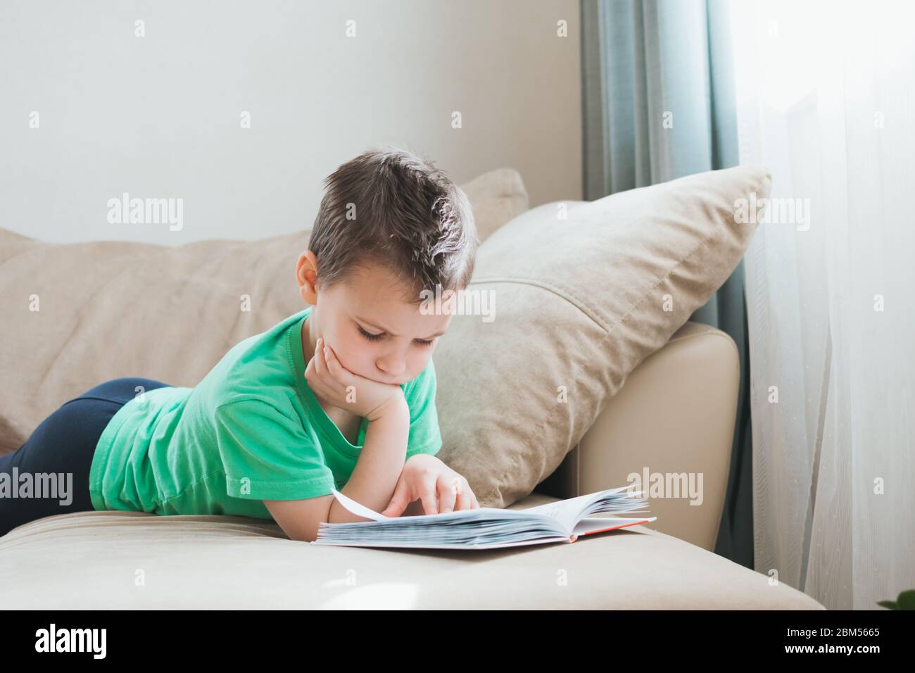 Ragazzo in maglietta verde leggendo un libro a casa sul divano. Formazione a distanza. Detox digitale. Ragazzo in quarantena rimanere a casa in isolamento auto. Foto Stock