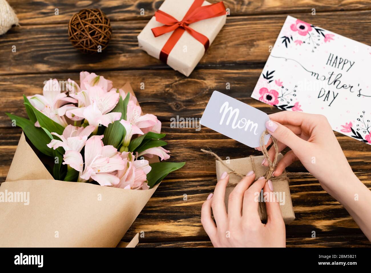 vista tagliata della donna che tocca tag con mamma scritta su scatola regalo vicino fiori rosa e biglietto di auguri con giorno felice madri su superficie di legno Foto Stock