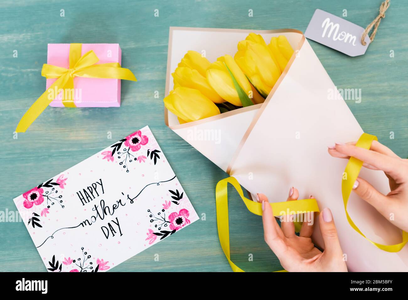 vista tagliata della donna che tocca il nastro vicino tulipani gialli, biglietto di auguri con le lettere di giorno delle madri felici e tag di mamma sulla superficie blu Foto Stock