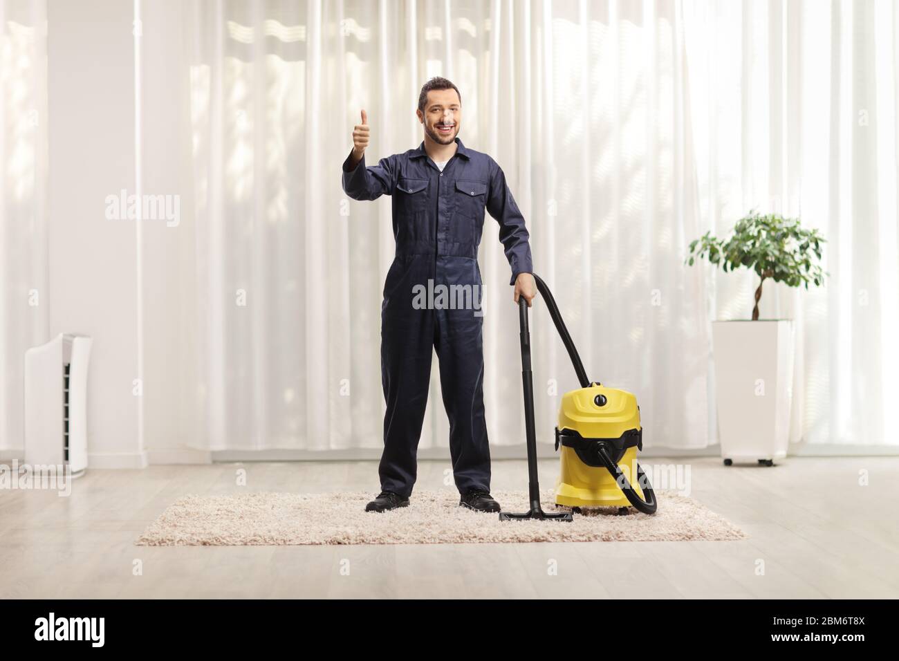 Ritratto a lunghezza intera di un lavoratore maschio da un cervice di pulizia con un hoover che pulisce una casa e mostra pollici in su Foto Stock
