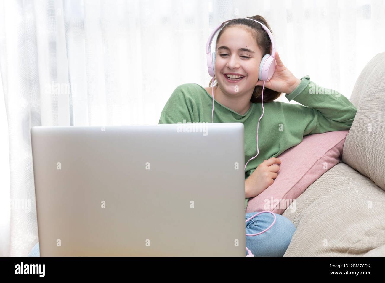 Ragazza seduta su un divano guardando un computer portatile con le cuffie mentre sorride in jeans e un pullover verde Foto Stock