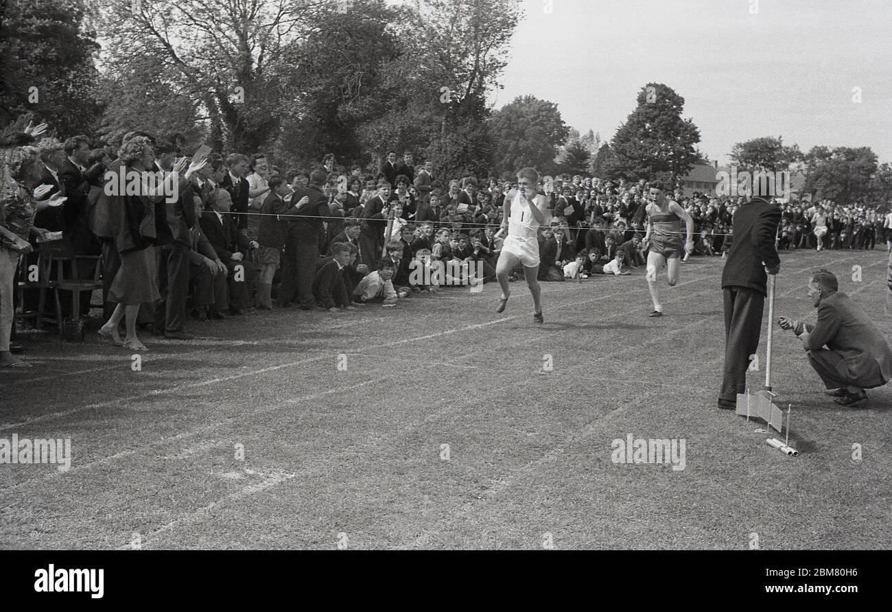 Scuola secondaria sport, Inghilterra, c1960, fuori su un percorso di erba, allietato da una grande folla di compagni di scuola e insegnanti, due giovani uomini che corrono duro per il nastro finale alla fine di una corsa di relè. Foto Stock