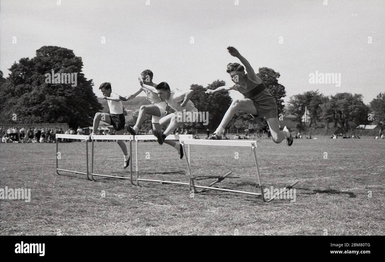 Giornata sportiva della scuola secondaria, Inghilterra, c1960s, pista e campo, quattro scolari che gareggiano all'esterno su un tracciato in erba in una corsa agli ostacoli, saltando oltre le barriere. Foto Stock