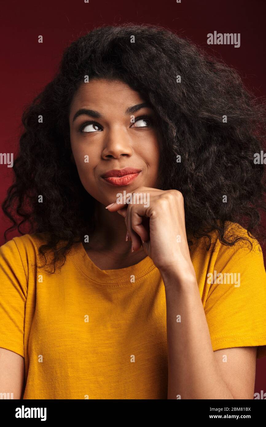 Immagine di bella donna marrone african american con capelli ricci che pensa e tocca il suo mento isolato su sfondo rosso Foto Stock