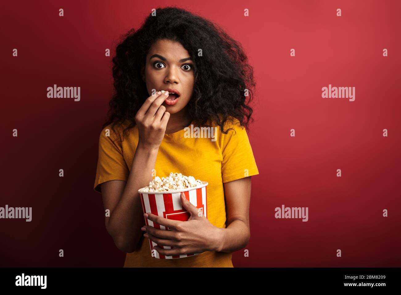 Immagine di una donna afro-americana scioccata brunette con capelli ricci che esprimono meraviglia mentre tenendo secchio popcorn isolato su sfondo rosso Foto Stock