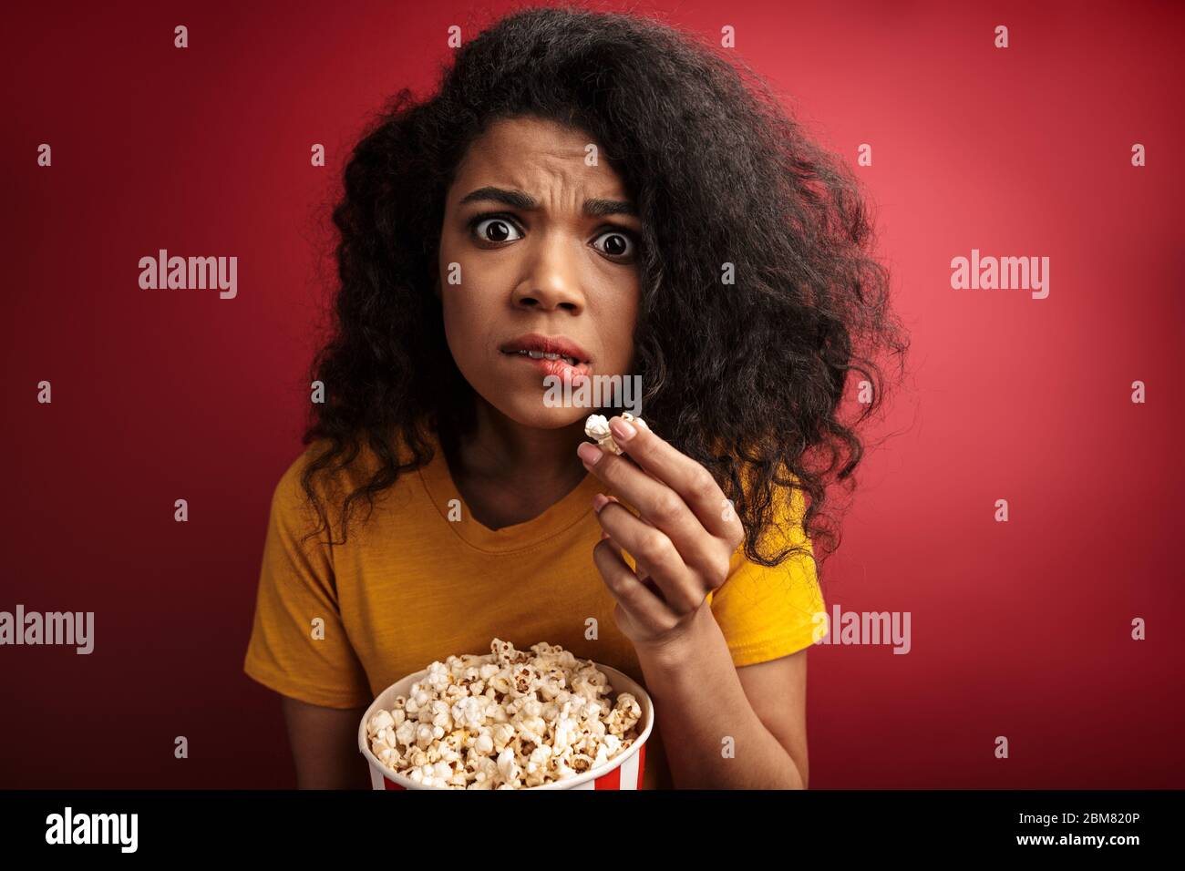 Immagine di una donna afro-americana scioccata brunette con capelli ricci che esprimono meraviglia mentre tenendo secchio popcorn isolato su sfondo rosso Foto Stock