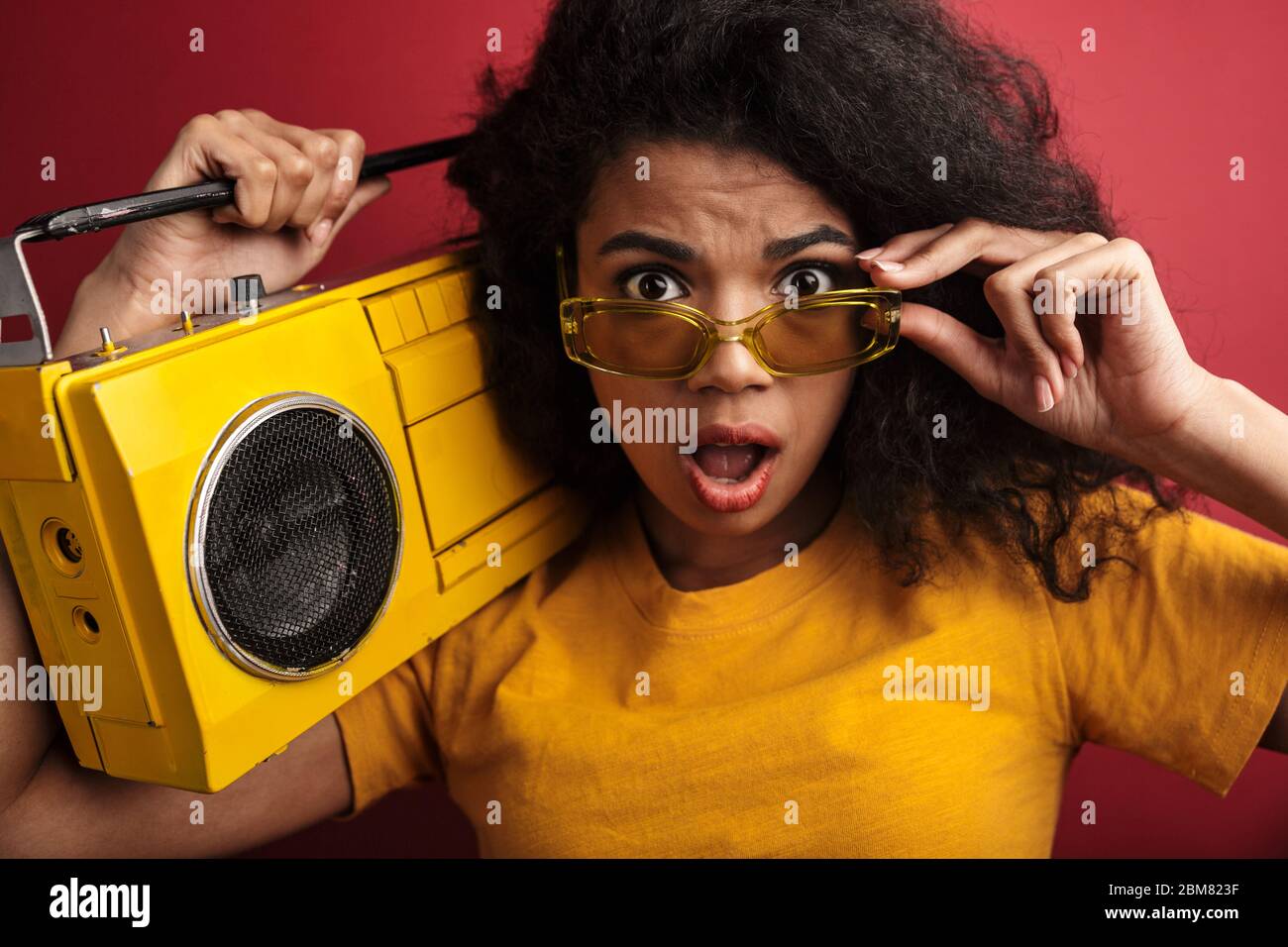 Immagine di una donna afro-americana bruna sorpresa con capelli ricci che tiene boombox vintage isolato su sfondo rosso Foto Stock