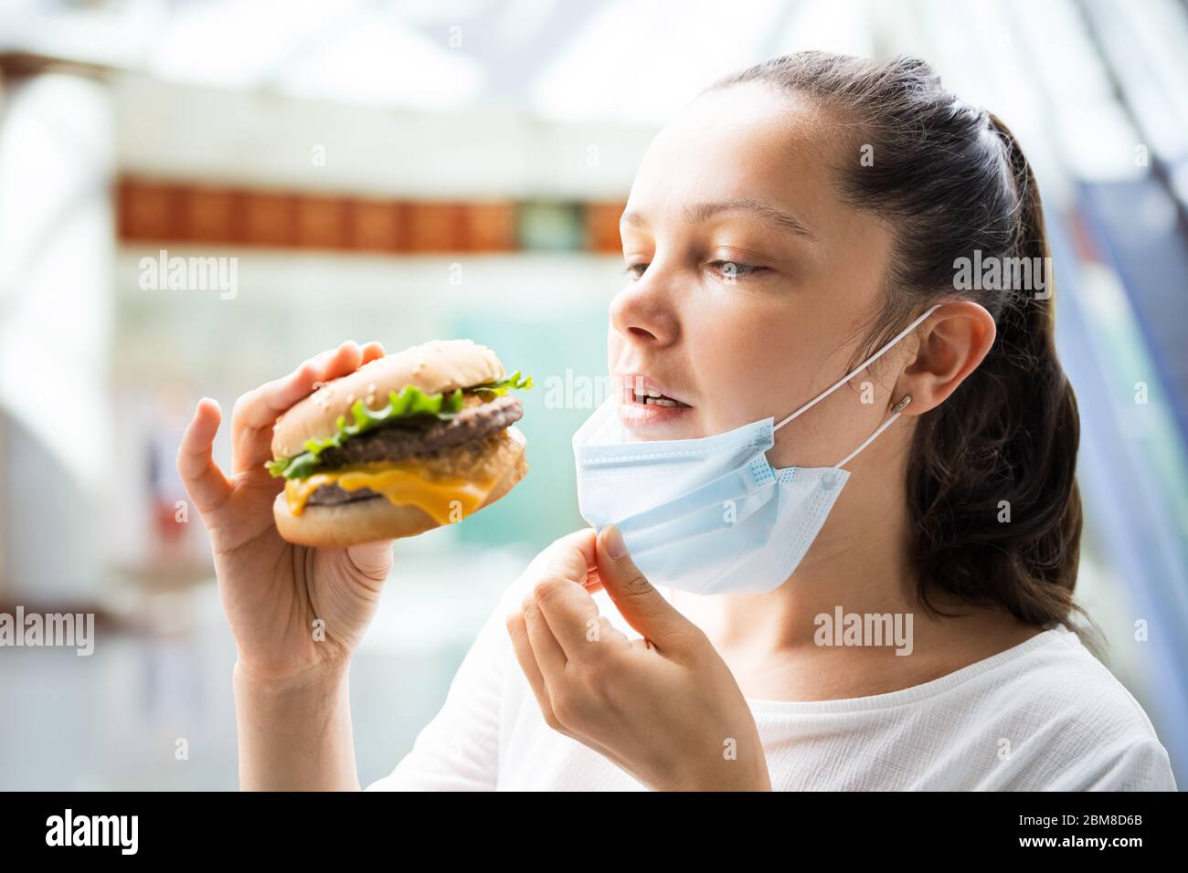 Donna che mangia hamburger di Fastfood in maschera facciale al ristorante Foto Stock