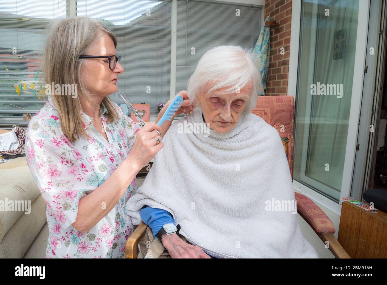 Covoid 19 restrizioni significa che la vecchia signora non può essere presa al parrucchiere come al solito, in modo che la sua caregiver visita rifinge e combatte i suoi capelli per lei come ge Foto Stock
