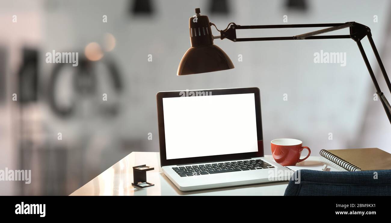 Computer portatile con schermo bianco vuoto posto su scrivania bianca e circondato da tazza da caffè, lampada, nota e penna. Spazio di lavoro ordinato. Photograp Foto Stock
