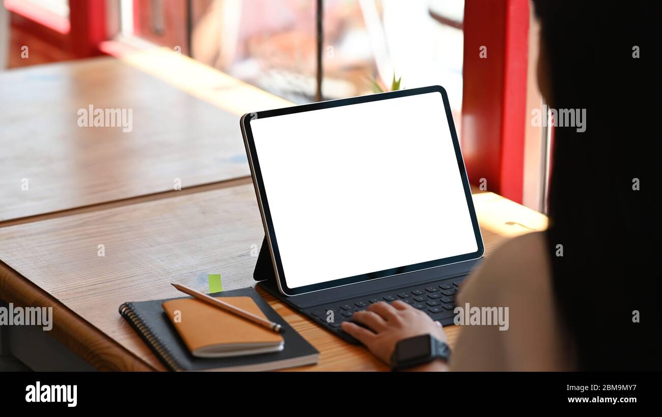 Immagine ritagliata delle mani creative della donna che battono su un tablet di computer bianco a schermo vuoto che mette su una scrivania in legno e circondato da notebook a. Foto Stock