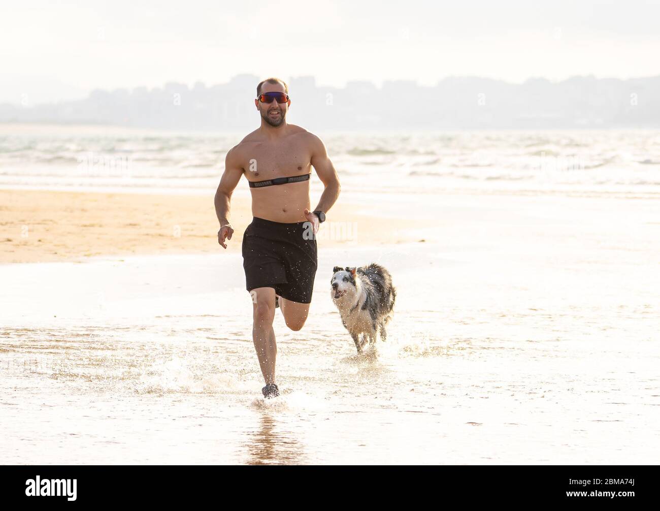 Uomo sportivo che corre con cani e allenamenti con cardiofrequietta sulla spiaggia adatta ai cani. Allenamenti all'aperto e jogging con il tuo migliore amico. Sano OW Foto Stock