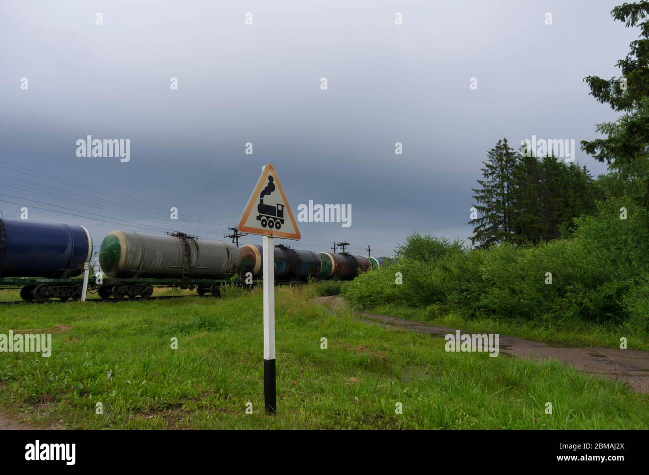 Cartello stradale 'attraversamento ferroviario senza barriera' sullo sfondo di un treno con serbatoi di petrolio e una strada di campagna Foto Stock