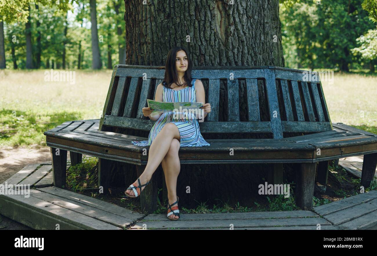 Una ragazza in una terrina seduta su una panchina del parco. Ragazza con una mappa turistica seduta su una panca di legno. Bella ragazza con una mappa turistica nel parco o Foto Stock