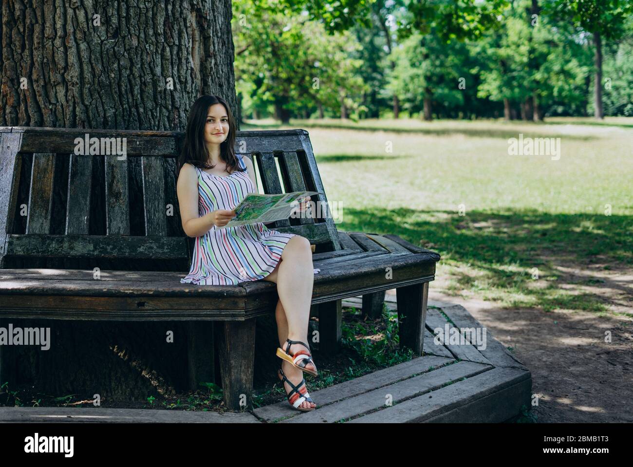 Una ragazza in una terrina seduta su una panchina del parco. Ragazza con una mappa turistica seduta su una panca di legno. Bella ragazza con una mappa turistica nel parco o Foto Stock